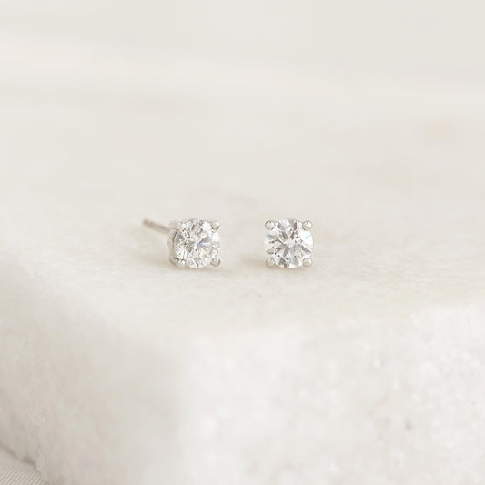 Two diamond stud earrings with White Gold settings are placed on a white textured surface. The image conveys elegance and simplicity.