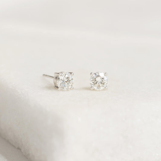 Two White Gold diamond stud earrings on a textured white surface, showcasing a minimalist and elegant design. The diamonds sparkle subtly in soft lighting.