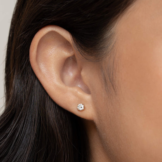 Close-up of a woman's ear wearing a small, sparkling diamond stud earring. The skin tone is warm, and the hairstyle is straight, dark hair.