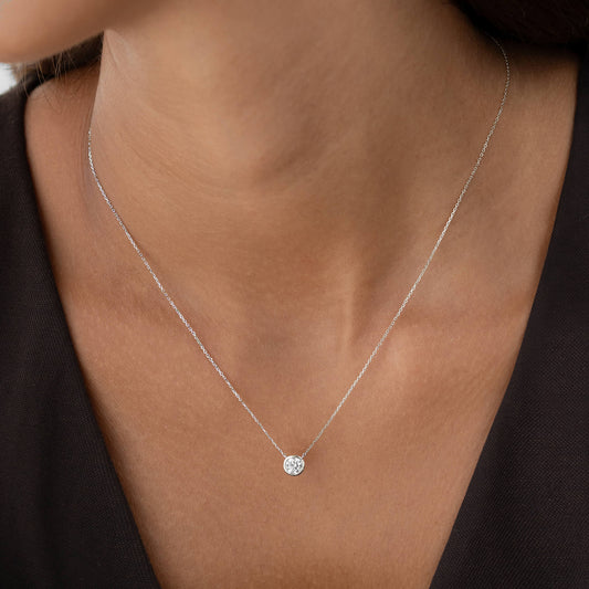 A close-up view of a person wearing a delicate White Gold chain necklace with a single, round diamond solitaire pendant held in a minimalist bezel setting, worn against a simple dark-colored top.