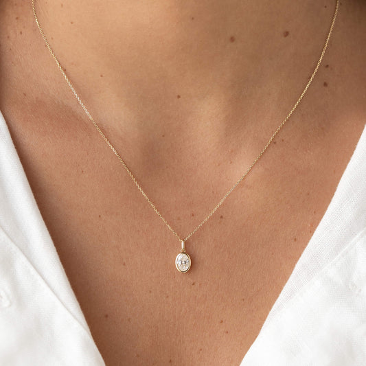Yellow Gold oval shaped pendant necklace with a delicate chain, worn centered on the chest over a white top.