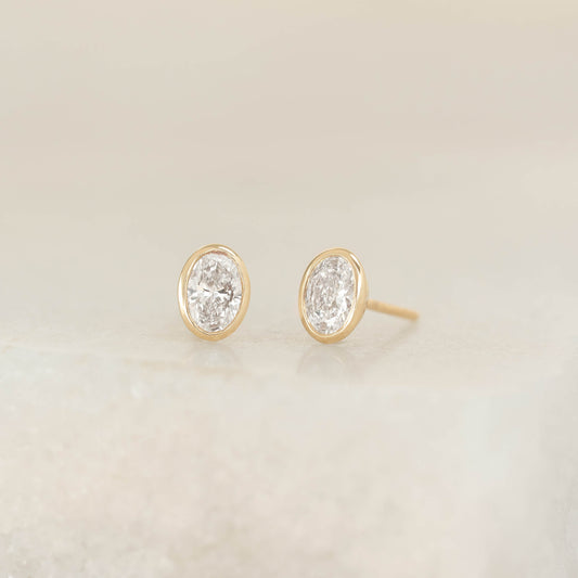 Pair of oval diamond stud earrings set in polished Yellow Gold bezels, displayed on a soft neutral surface.