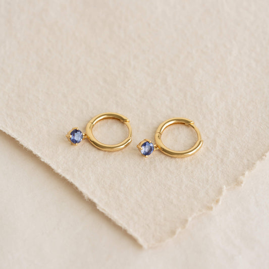Pair of small Gold hoop earrings with dangling oval light blue gemstones, placed on a soft, textured off-white fabric surface.