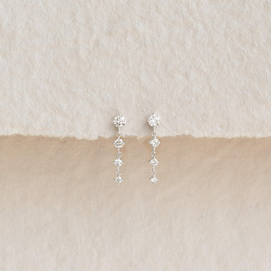 Pair of delicate drop earrings featuring sparkling diamond-shaped stones arranged in a vertical design, displayed on a textured beige and cream background.
