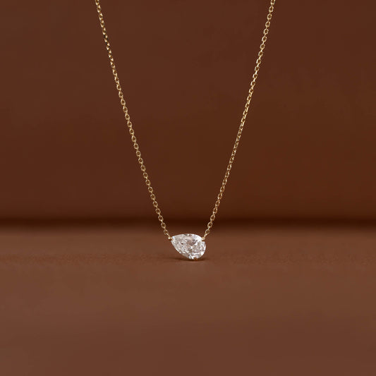 Close-up of a Gold chain necklace with a brilliant pear-shaped clear gemstone pendant displayed against a warm brown background.