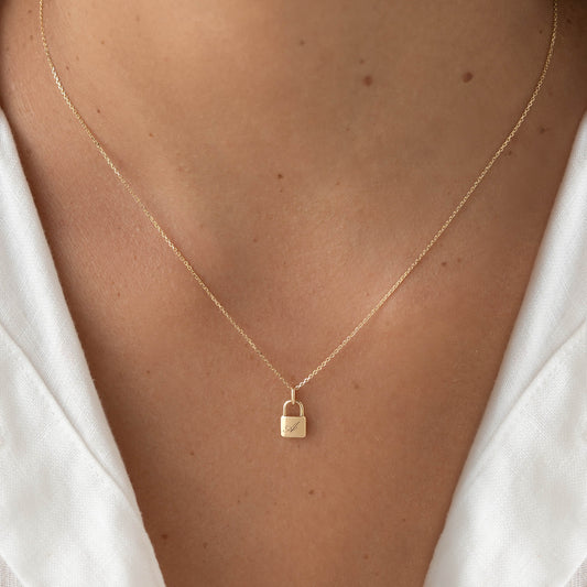 Yellow Gold padlock shaped pendant necklace with a delicate chain, worn centered on the chest over a white top.