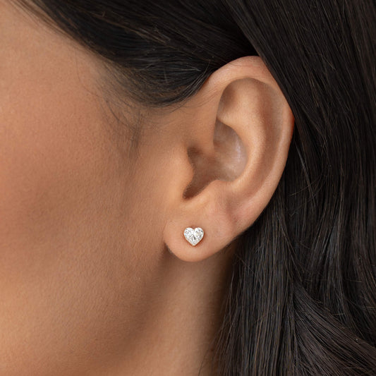 Close-up of a person’s ear wearing a small Gold heart-shaped diamond stud earring, with smooth skin and dark hair tucked behind the ear.