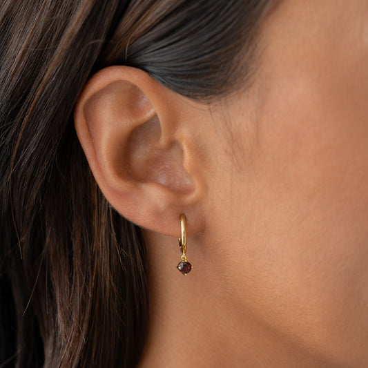 Close-up of a gold vermeil January Birthstone Drop Huggie Earrings - Garnet worn on a woman’s ear against neutral background