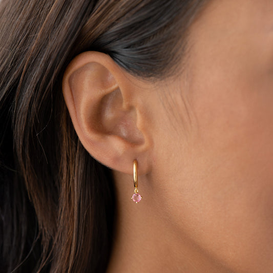 Close-up of an ear wearing July Birthstone Drop Huggie Earrings - Pink Tourmaline with gold vermeil setting on neutral background