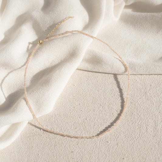 Sunstone Necklace with delicate peach-colored beads displayed on a textured light background with draped fabric