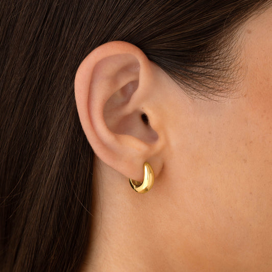 Close-up of an ear wearing gold Dome Huggie Earrings - Caja with a tapered dome shape in soft natural light