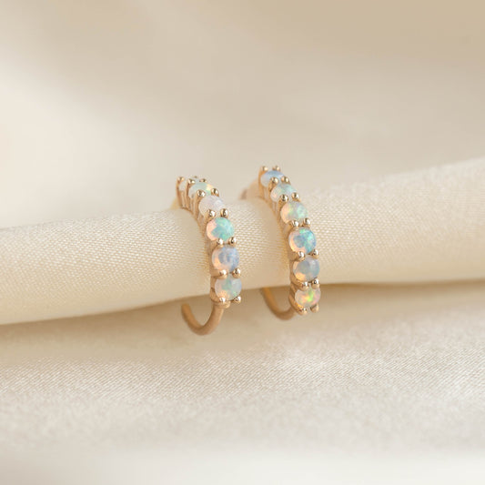 Close-up of Opal Huggie Earrings 14k Gold - Calypso featuring pave set iridescent opal gemstones on a soft beige fabric background