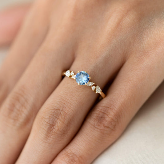 The central, round brilliant-cut sapphire displays a vivid light blue hue, secured within six prominent Gold prongs.