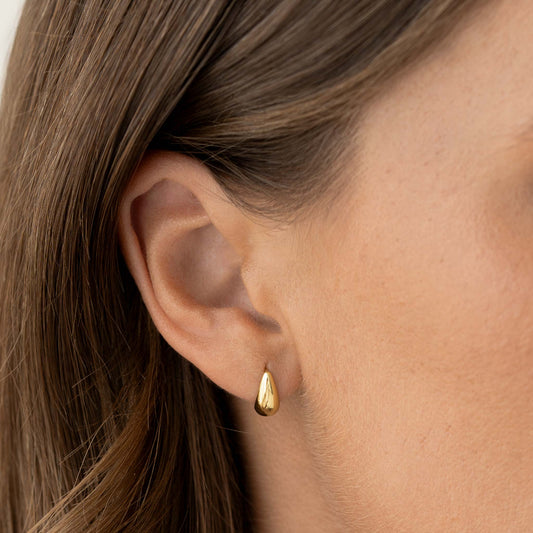 Close-up of a woman wearing Tapered Dome Huggie Earrings - Brianne in gold vermeil on a neutral background