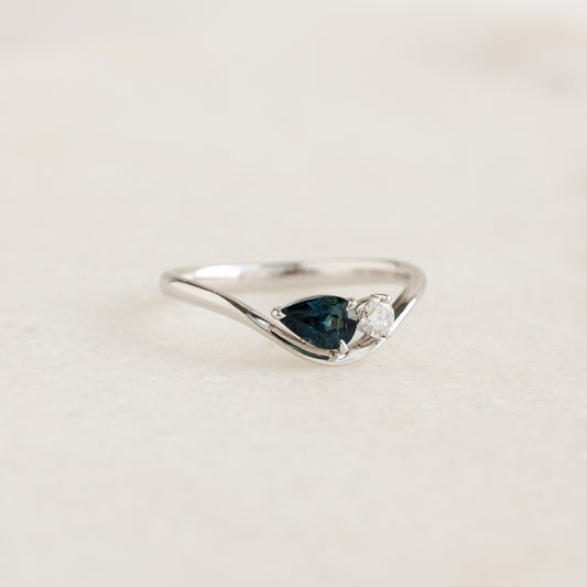 The White Gold ring features a sleek, asymmetrical wavy band that dips gracefully down to secure a horizontally-set, pear-cut deep teal sapphire.