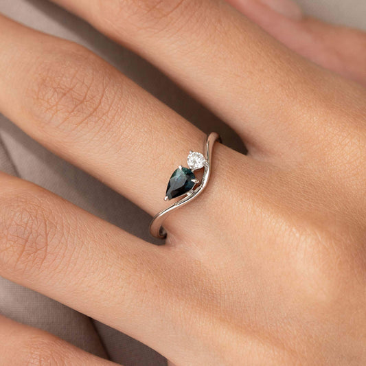 The photo focuses closely on the hand, offering a clear view of how the minimalist, modern jewelry piece sits on the finger against a softly blurred, neutral background.