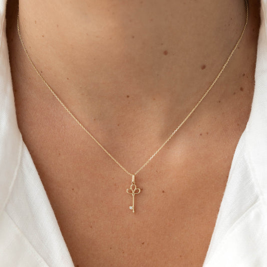 Yellow Gold key shaped pendant necklace with a delicate chain, worn centered on the chest over a white top.