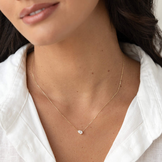 Close-up of a woman wearing a delicate Gold chain necklace with a small pear diamond pendant, styled with an open white shirt against a soft neutral background.