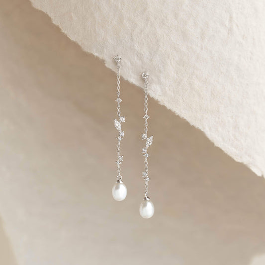 Delicate silver drop earrings featuring sparkling leaf-shaped details and elegant white pearl pendants, displayed against a soft cream textured background.