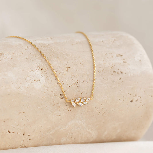 Gold necklace with a delicate leaf pendant adorned with small diamonds, elegantly displayed on a textured beige stone. The tone is elegant and refined.