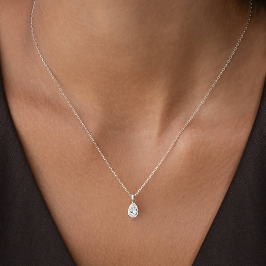A close-up shot of a person's neckline wearing a delicate, White Gold chain necklace. The necklace features a small, sparkling, pear-shaped diamond pendant that rests gently against the skin.