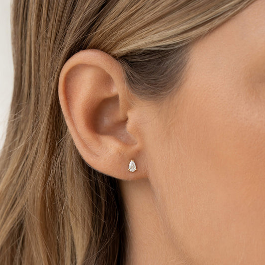 Close-up of a person's ear wearing a Gold small teardrop-shaped diamond earring. The image conveys elegance and simplicity, with blonde hair framing the ear.
