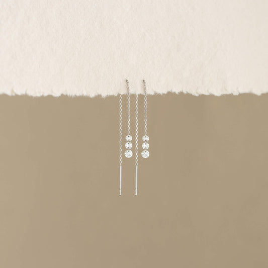 White Gold dangling earrings with sparkling beads hang against a torn paper edge and soft beige background, conveying elegance and simplicity.