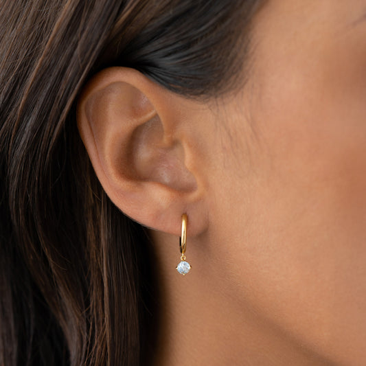 Close-up of a woman’s ear wearing March Birthstone Drop Huggie Earrings - Aquamarine with gold vermeil finish on neutral background