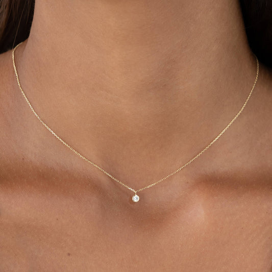 A close-up shot of a person wearing a very delicate Gold necklace with a tiny, round brilliant diamond solitaire pendant set in a minimalist bezel setting.