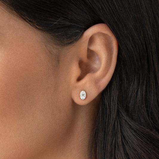Close-up of a Woman's ear wearing a small Gold oval diamond stud earring set in Gold metal, with smooth skin and dark hair tucked behind the ear.