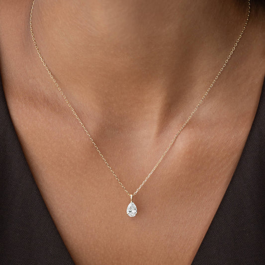 A close-up shot of a person's neckline wearing a delicate, Gold chain necklace. The necklace features a small, sparkling, pear-shaped diamond pendant that rests gently against the skin.