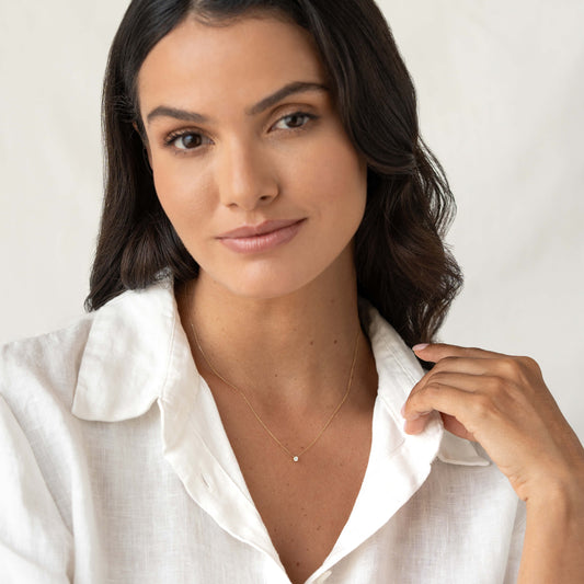 Woman with long dark wavy hair wearing a white collared blouse and a delicate Gold necklace with a small round gemstone pendant. | 0.15 ct tw