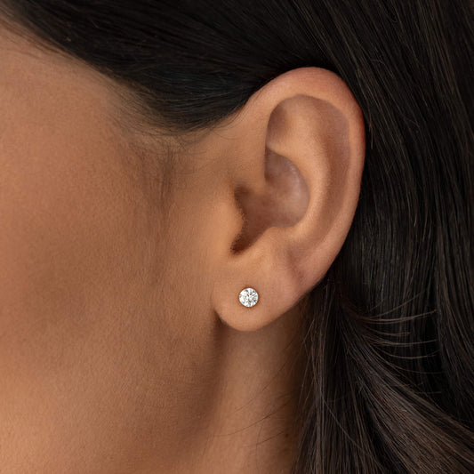 A close-up of a person's ear wearing a small round stud earring with a clear gemstone in a Gold setting. | 0.5 ct tw