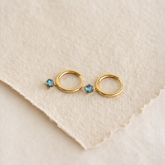 Close-up of September Birthstone Drop Huggie Earrings - London Blue Topaz with gold vermeil setting on textured paper background