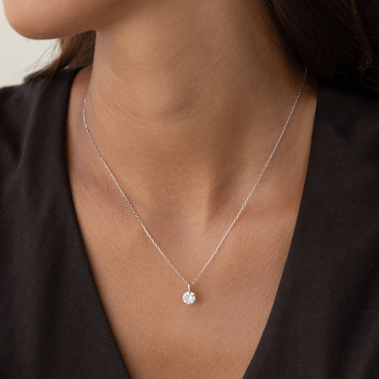 A portrait of a woman with dark hair wearing a delicate White Gold chain necklace with a small round brilliant diamond solitaire pendant held in a prong setting, paired with a dark V-neck top.