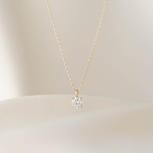 A close-up shot of a delicate Gold necklace with a small, round diamond solitaire pendant in a prong setting, draped over a textured, light beige surface.