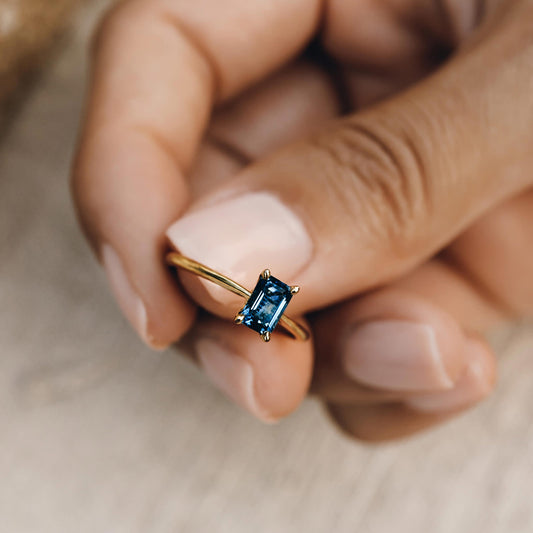 Close-up of a hand holding the Baguette Ring London Blue Topaz - Charlotte with a gold band and rectangular gemstone