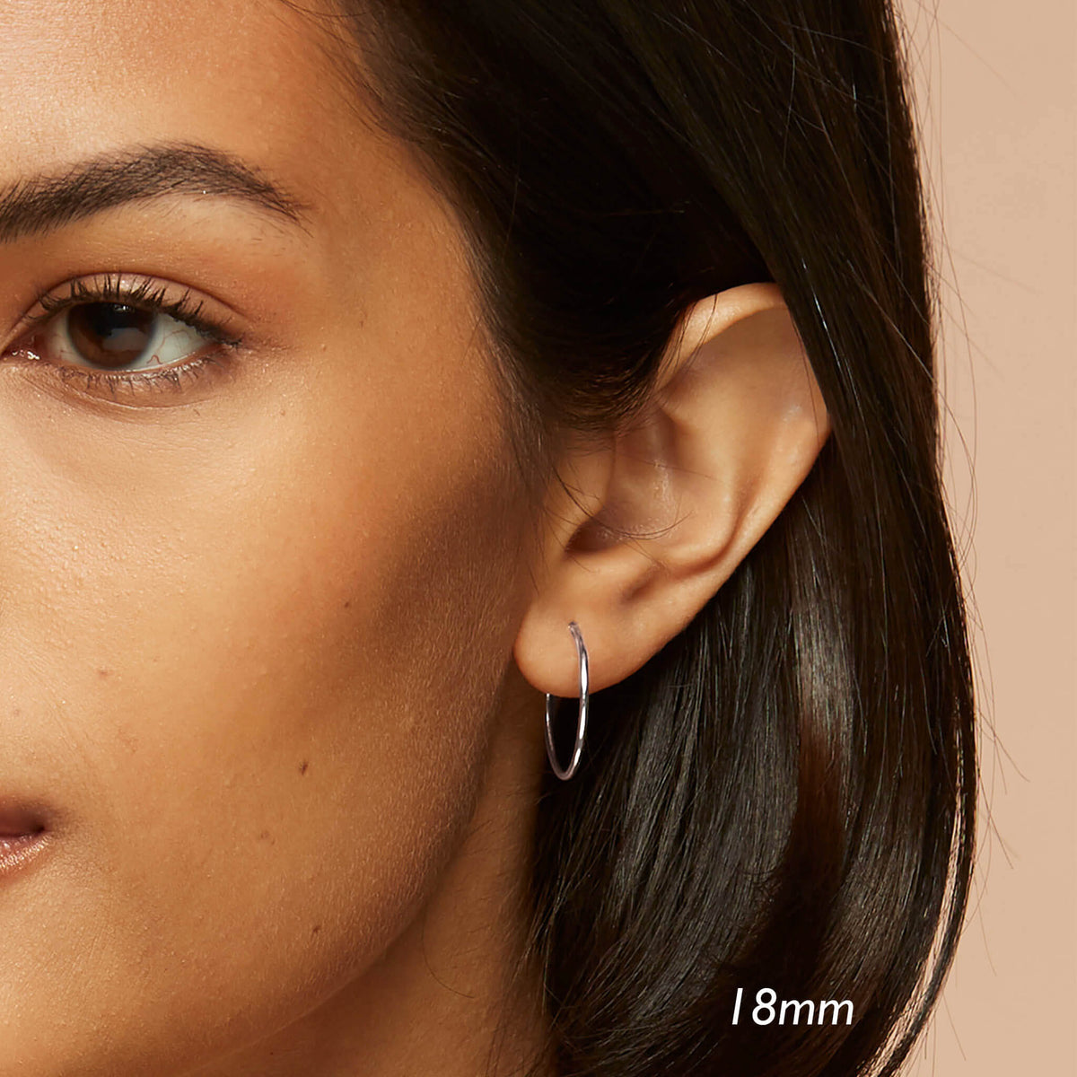 Close-up of a woman wearing White Gold Hoops 18mm - Sonia with smooth skin and dark hair against a beige background