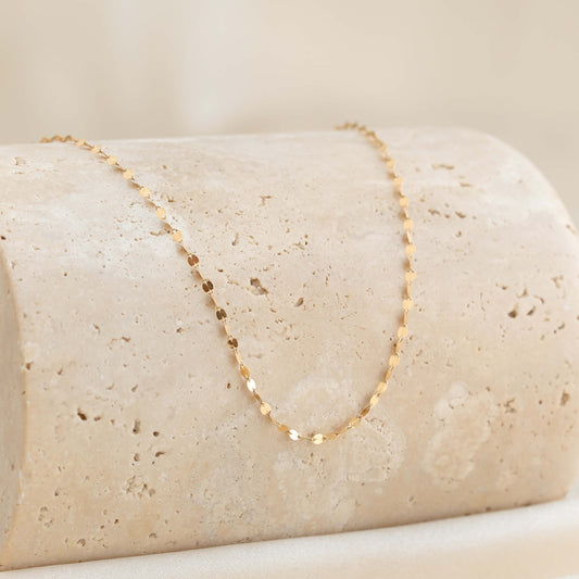 A delicate Gold chain necklace with small, round links elegantly draped over a textured beige stone surface, conveying simplicity and elegance.