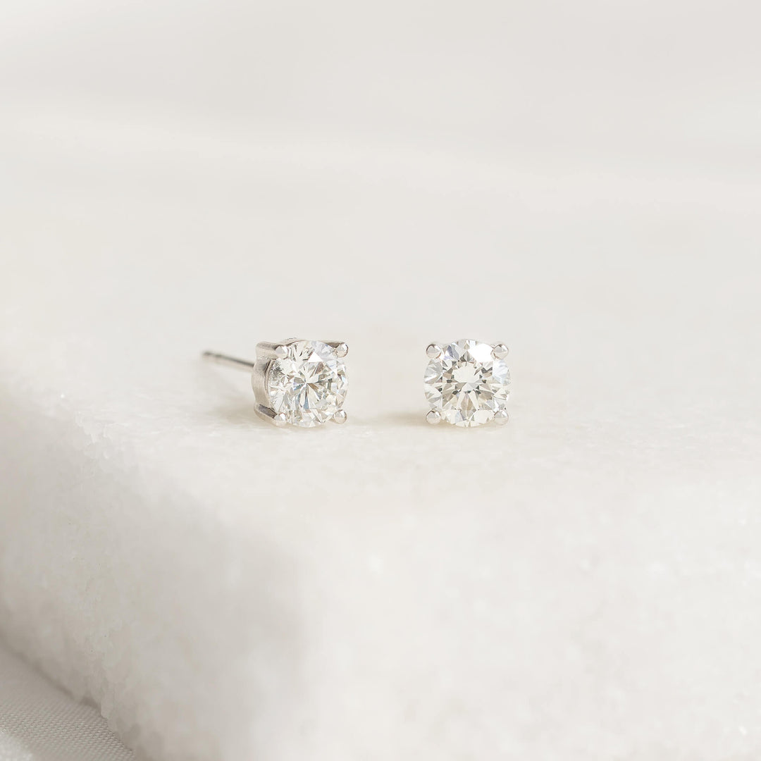 Two White Gold diamond stud earrings on a textured white surface, showcasing a minimalist and elegant design. The diamonds sparkle subtly in soft lighting.