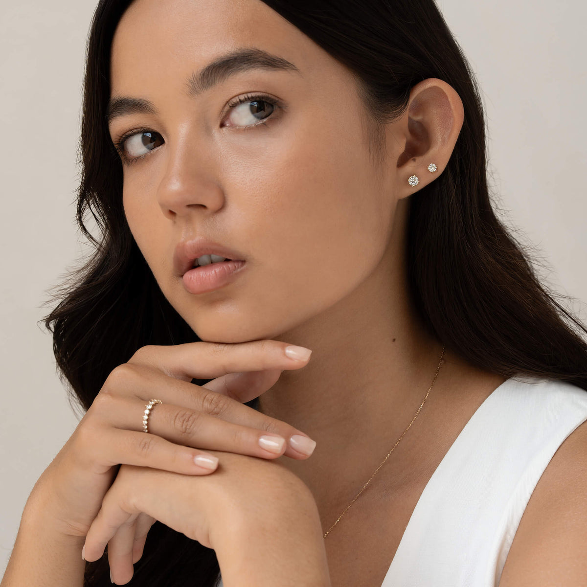 A woman with long dark hair gazes thoughtfully to the side, wearing a white top and minimalistic Gold jewelry, including stud earrings and a delicate ring.