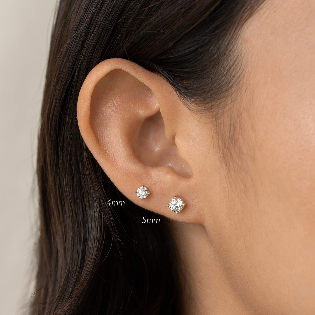 Close-up of a woman's ear wearing two diamond stud earrings. The top earring is 4mm, and the bottom is 5mm. The image has a neutral tone.