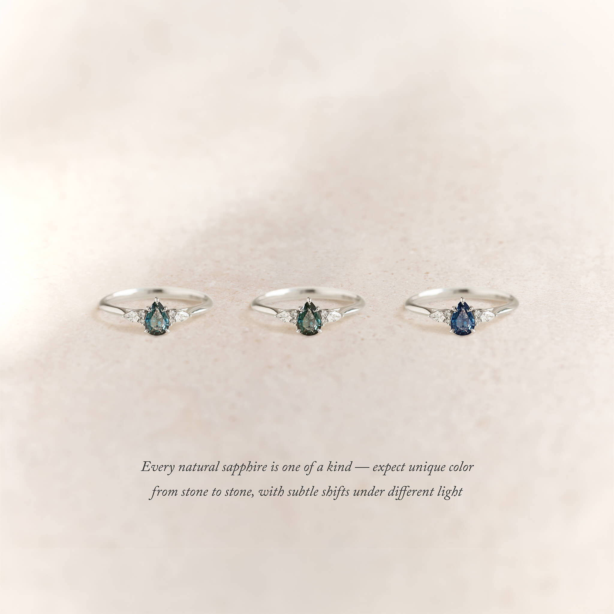Three White Gold rings with pear-shaped sapphires in varying hues are arranged side by side on a soft beige background.