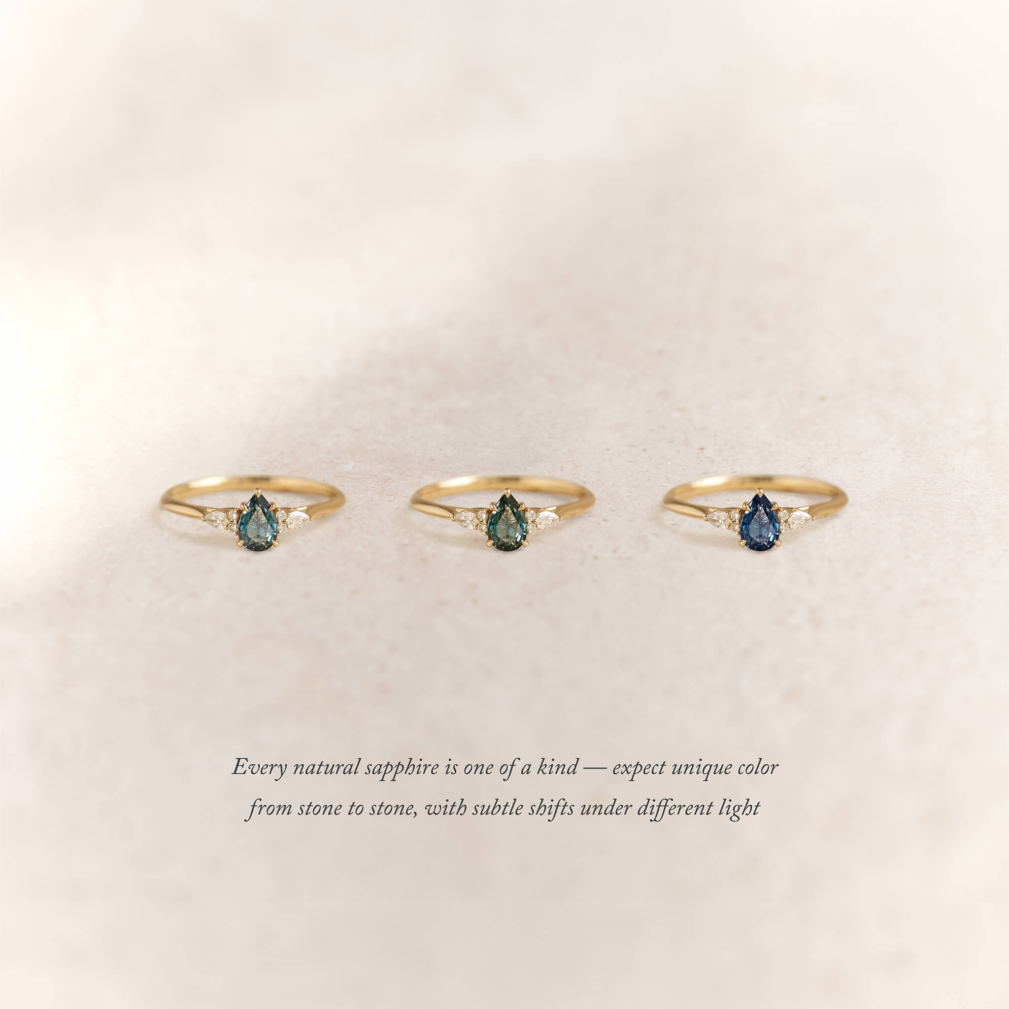 Three Gold rings with pear-shaped sapphires in varying hues are arranged side by side on a soft beige background.