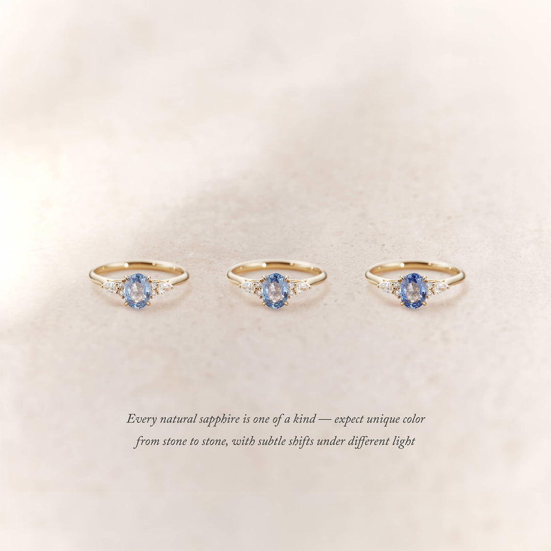 Three Yellow Gold rings with oval light blue sapphires and small diamond accents on each side are displayed side by side on a soft beige background.
