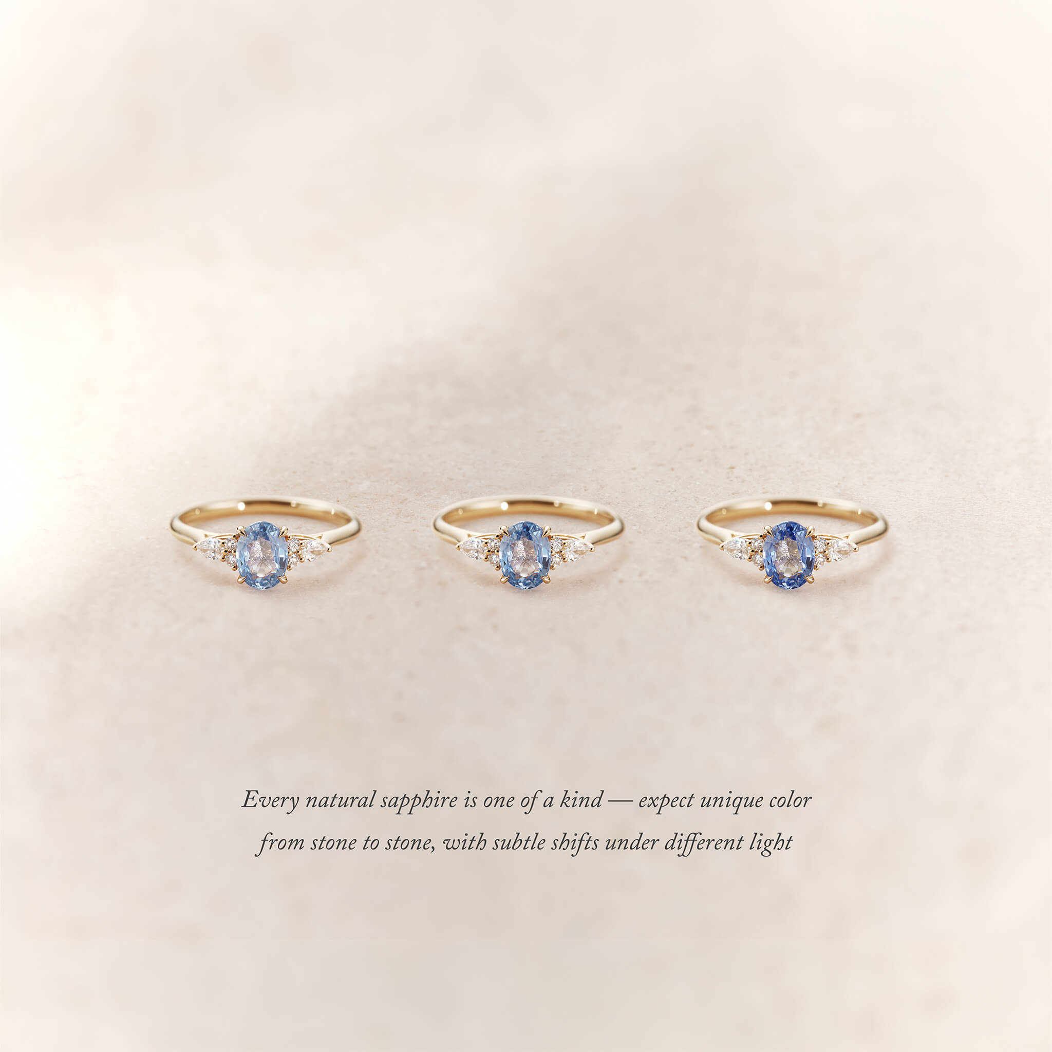 Three Yellow Gold rings with oval light blue sapphires and small diamond accents on each side are displayed side by side on a soft beige background.