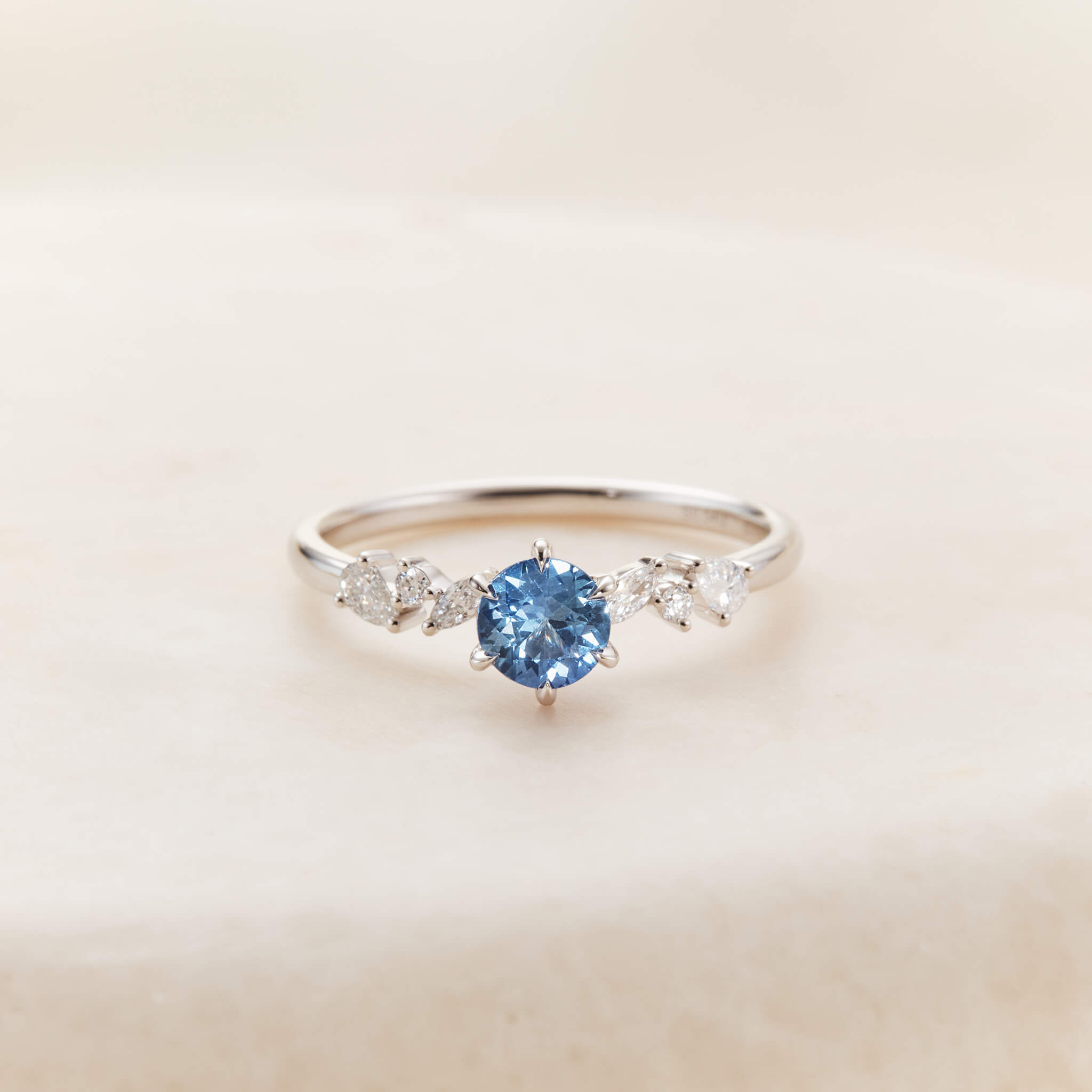 A close-up product photograph displays a delicate 14k White Gold ring centered against a textured, light beige stone surface.