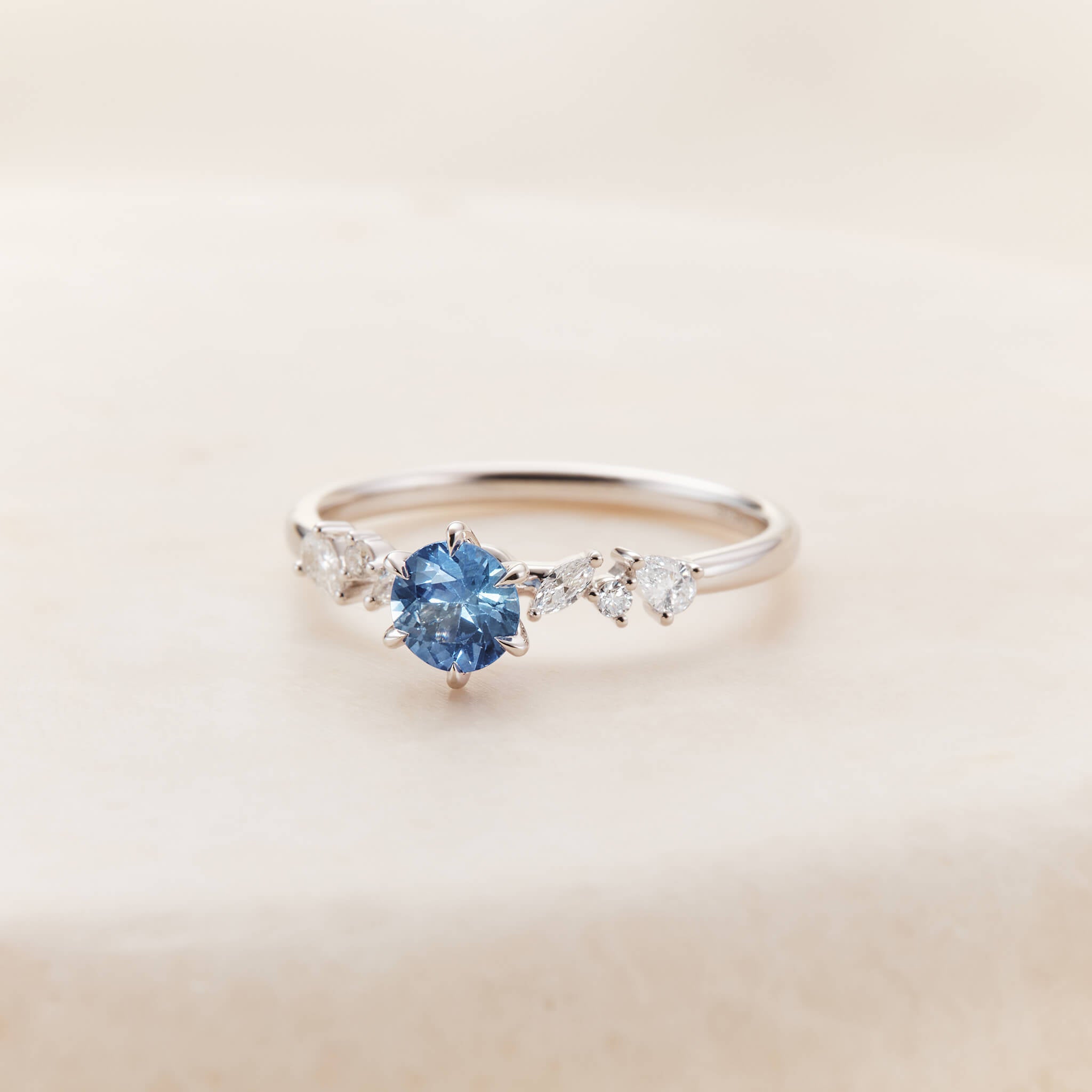 A close-up studio photograph showcases a delicate White Gold ring resting angled forward on a textured, pale beige stone surface.