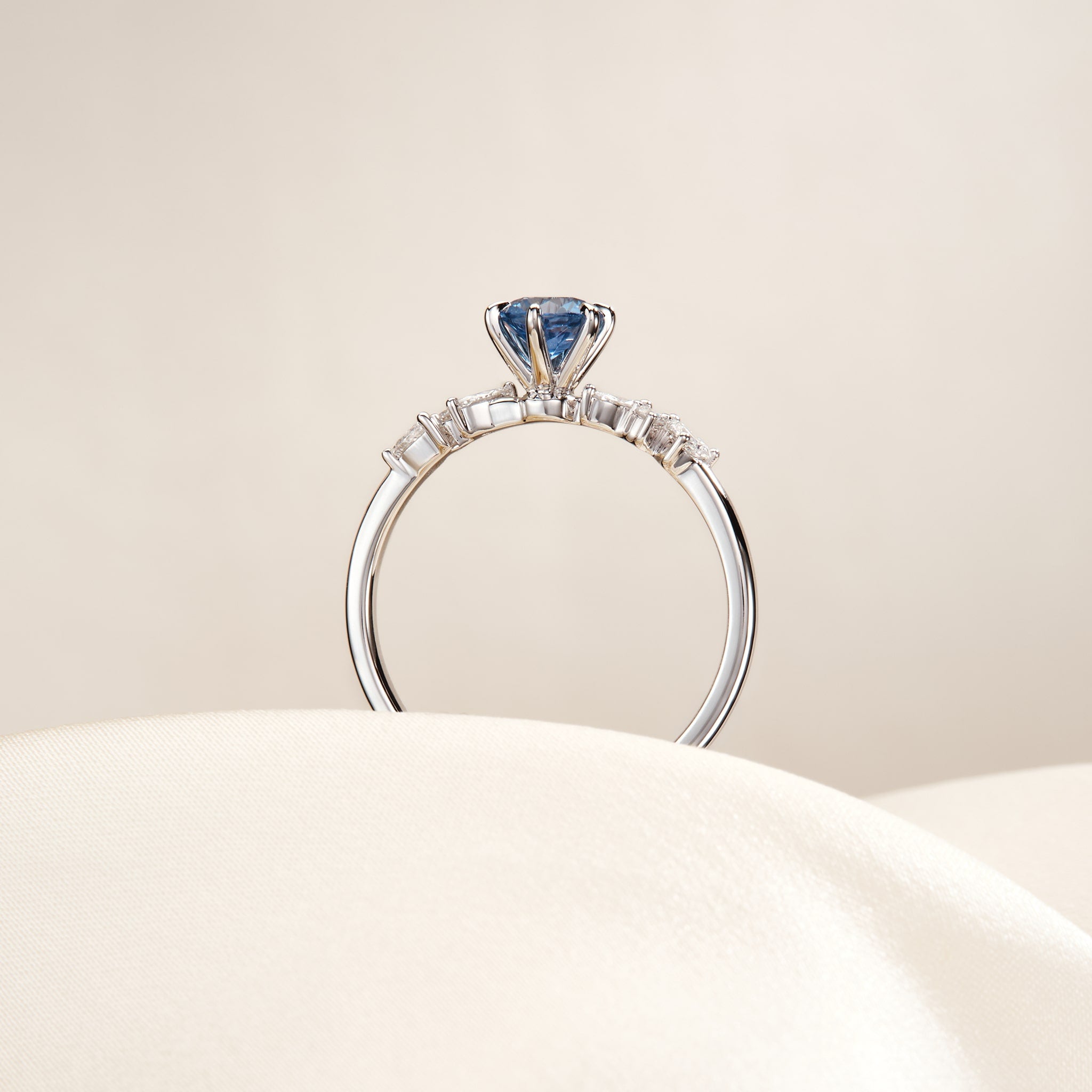 A studio product photograph showcases a delicate White Gold engagement ring resting upright and angled slightly forward on a smooth, pale cream stone surface.