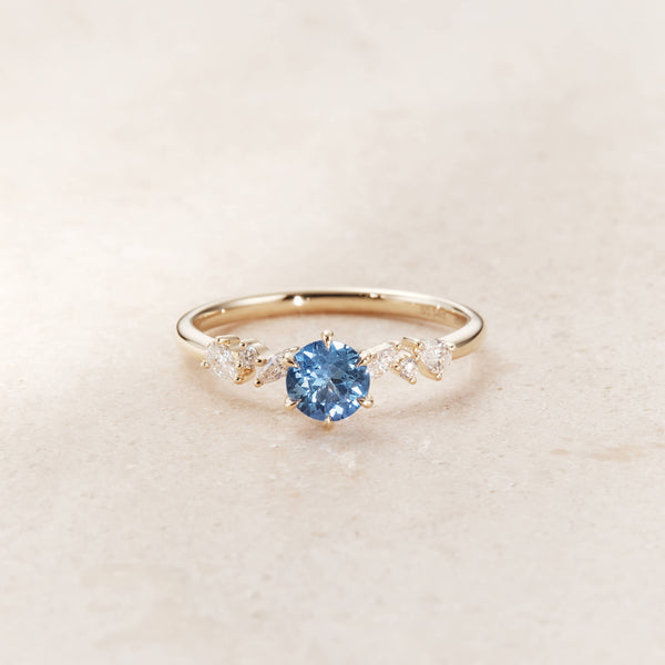 On each side of the central stone, the slender Gold band is accented by a small, symmetric cluster of sparkling white diamonds that form a subtle floral motif.
